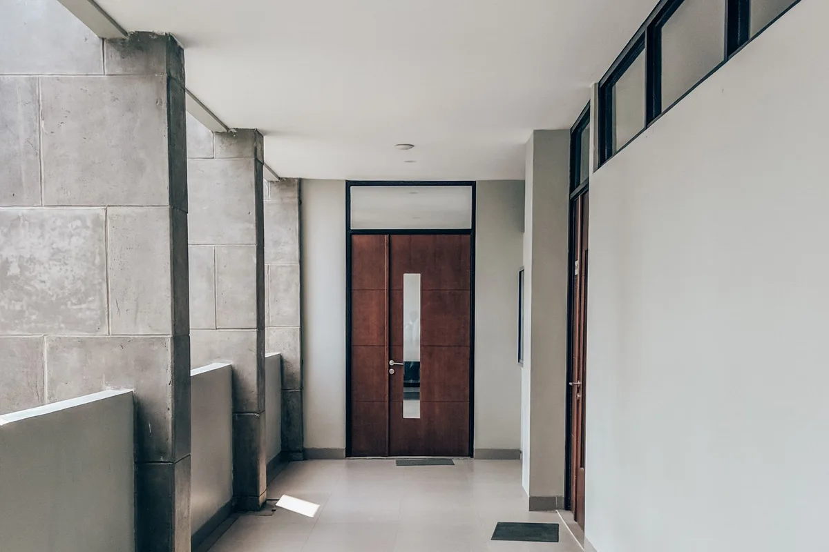 A clean, sunlit apartment corridor with a tiled floor leading to a wooden flat door with doormats outside it.