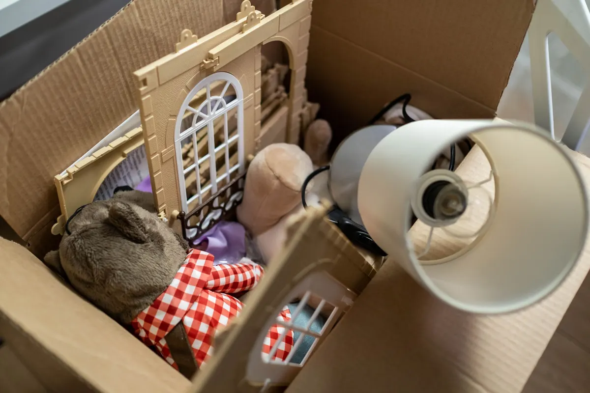 An open cardboard box packed with outgrown toys, a soft toy and a discarded lampshade ready to be taken away.
