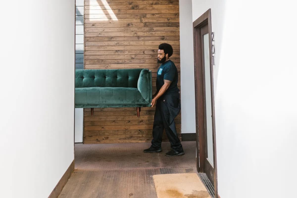 A mover carrying a green two-seater sofa along a narrow building corridor towards the exit.