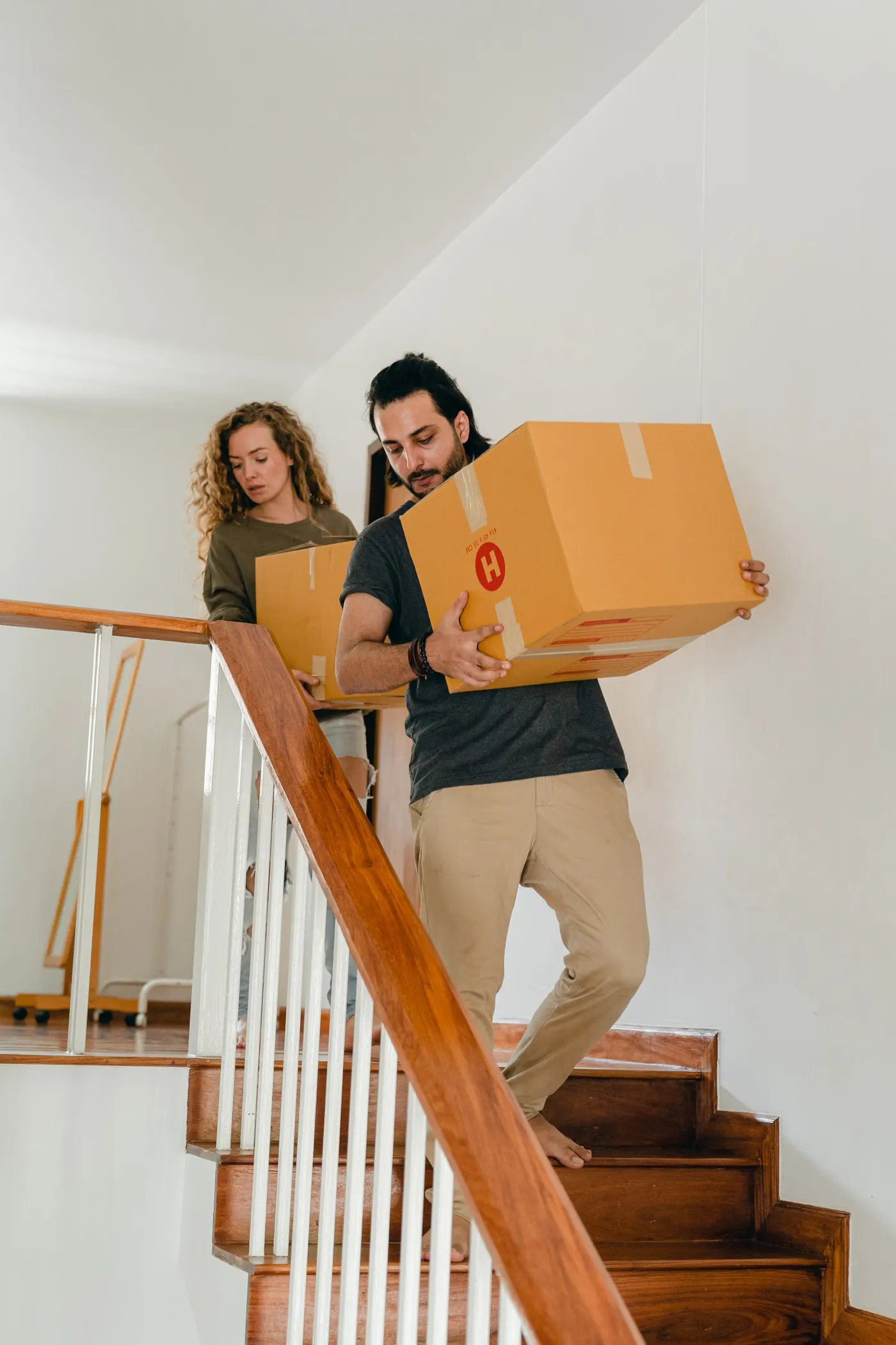 Two people carrying large cardboard moving boxes down an internal staircase inside a building.