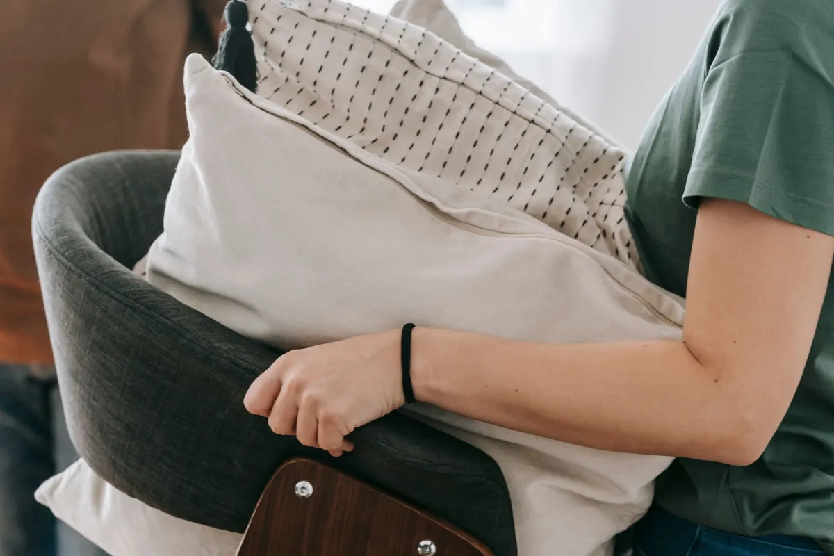 A person carrying a single wooden-framed armchair stacked with cushions across a room.