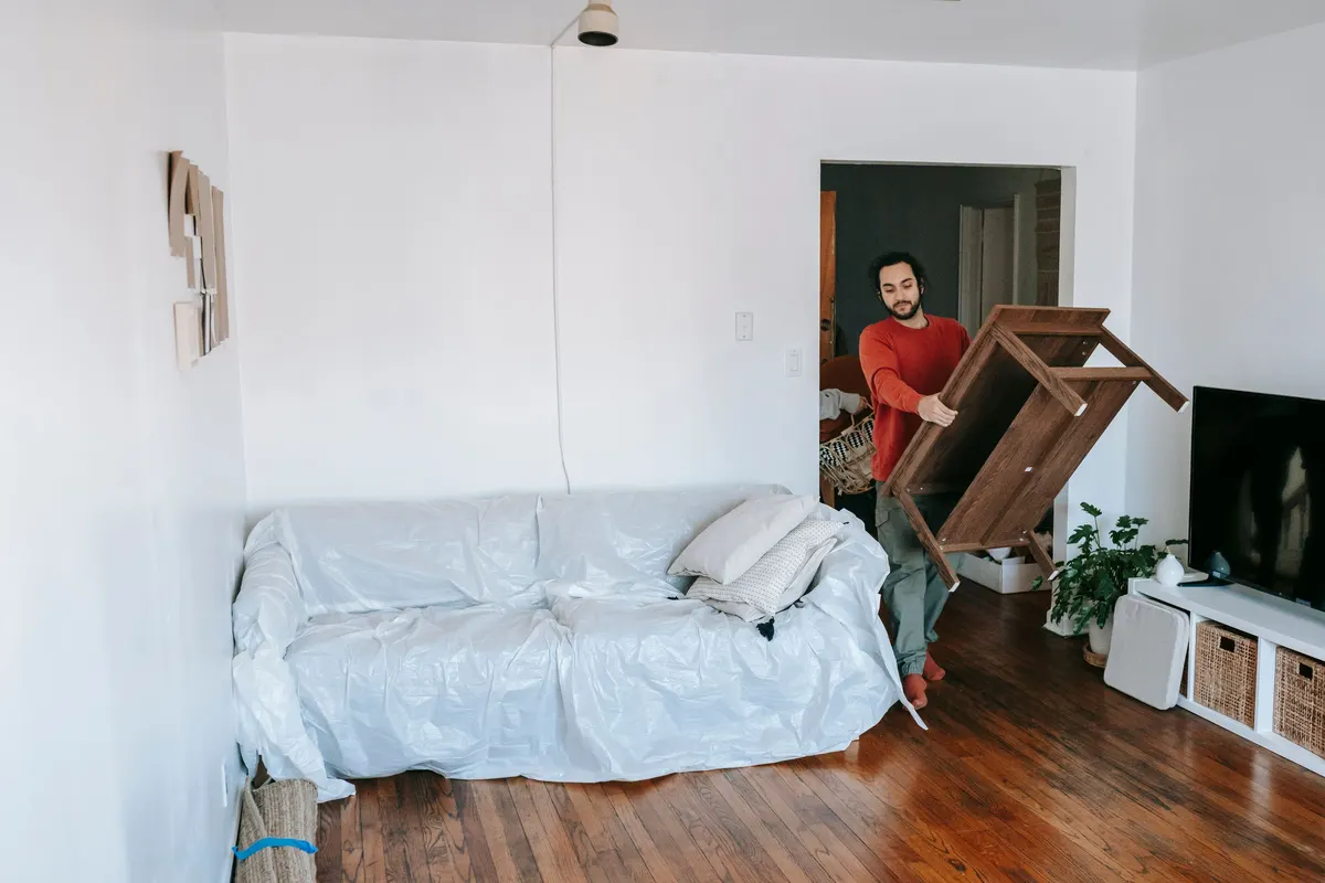 A man carrying a small wooden side table out of a living room past a sofa covered with a protective sheet.