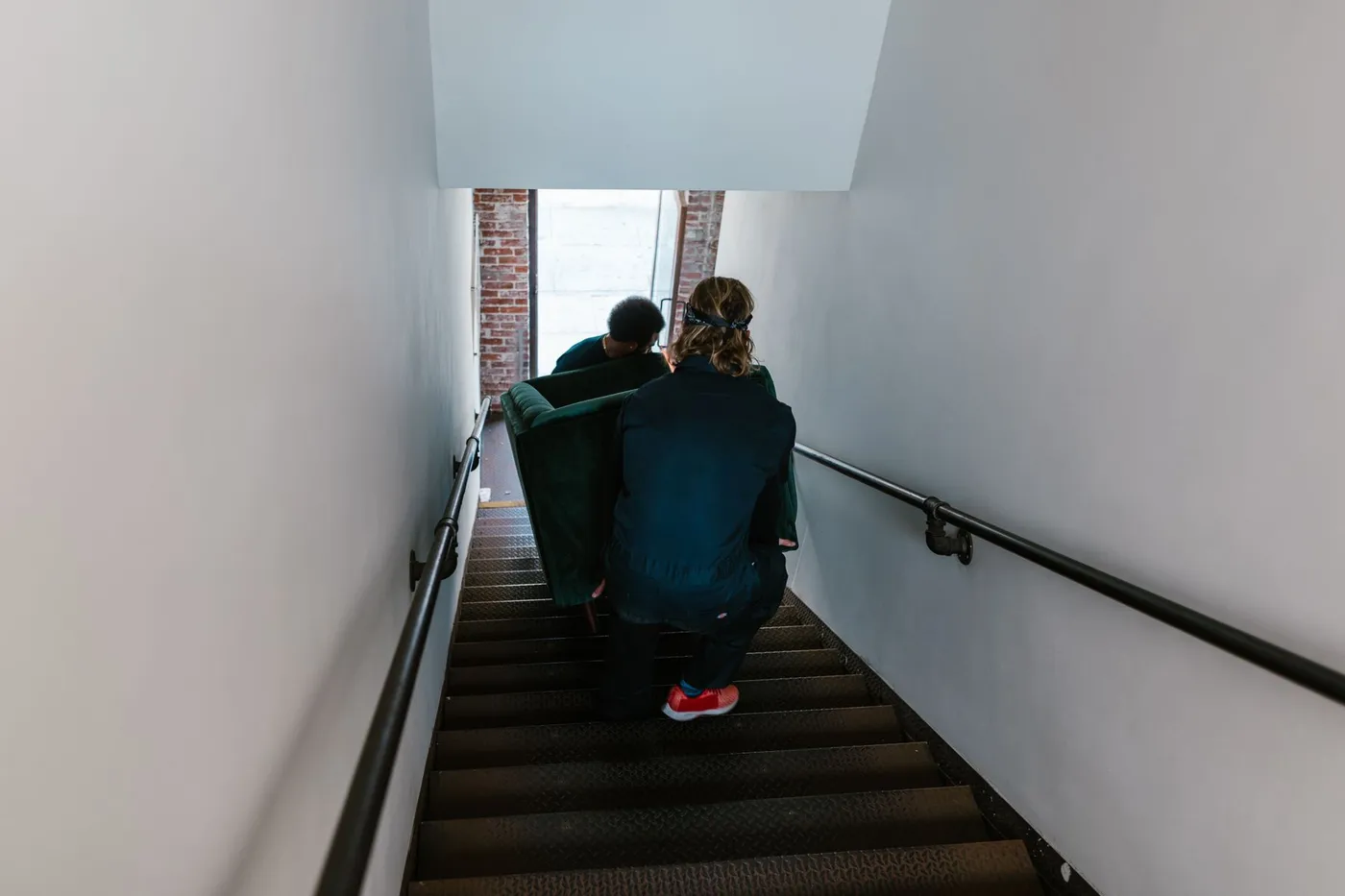 Two workers in overalls carrying a green sofa down a narrow indoor stairwell, viewed from the landing above.