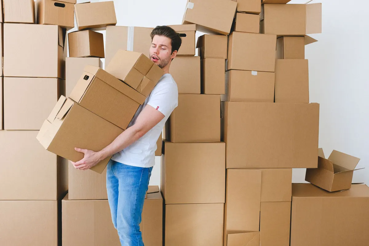 A man carrying a tall stack of cardboard boxes in front of a wall of stacked empty cartons.