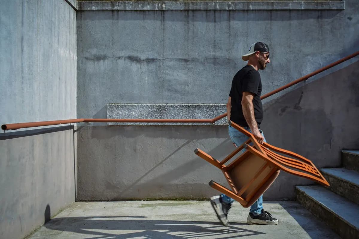 A man in a black T-shirt carrying a wooden chair upside down across a concrete landing beside a flight of steps.