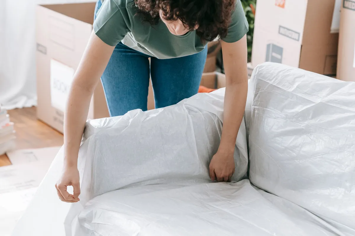 A woman smoothing a white protective sheet over a sofa in a room stacked with packed cardboard boxes.