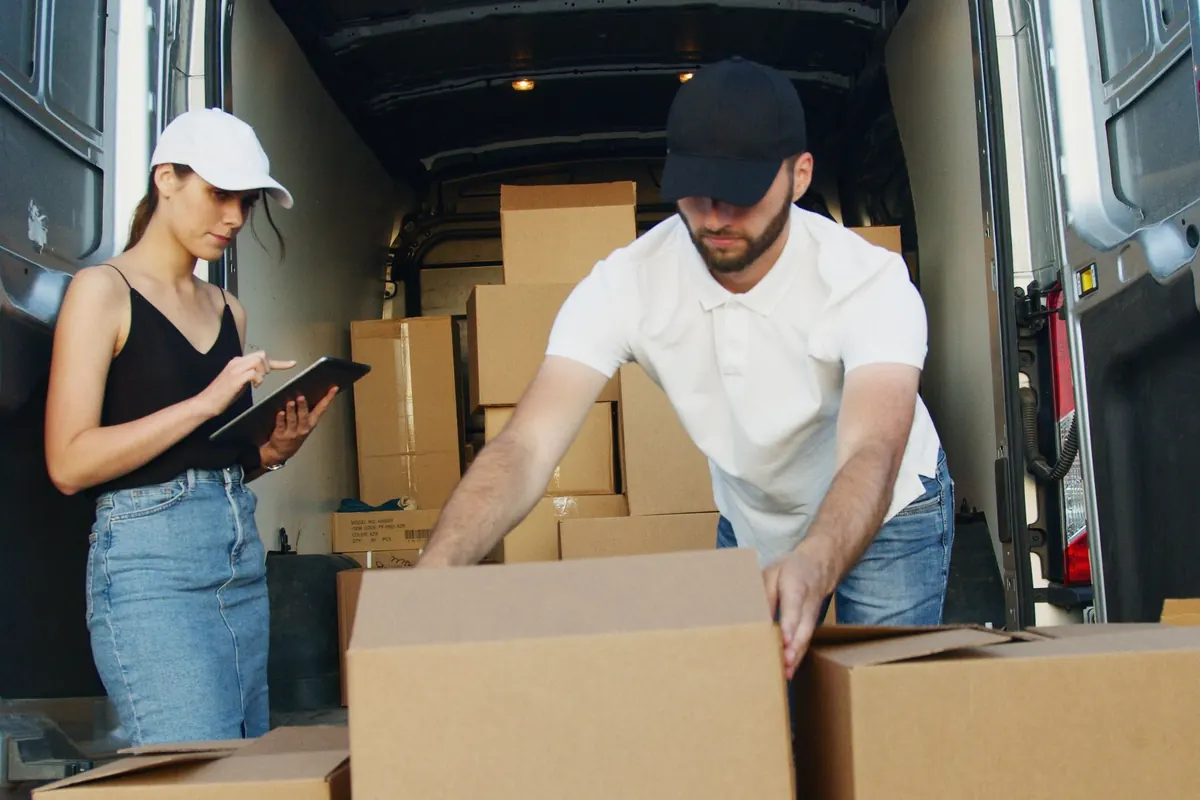 A worker in a white polo shirt stacking cardboard cartons at the open rear doors of a van while a colleague notes details on a tablet.