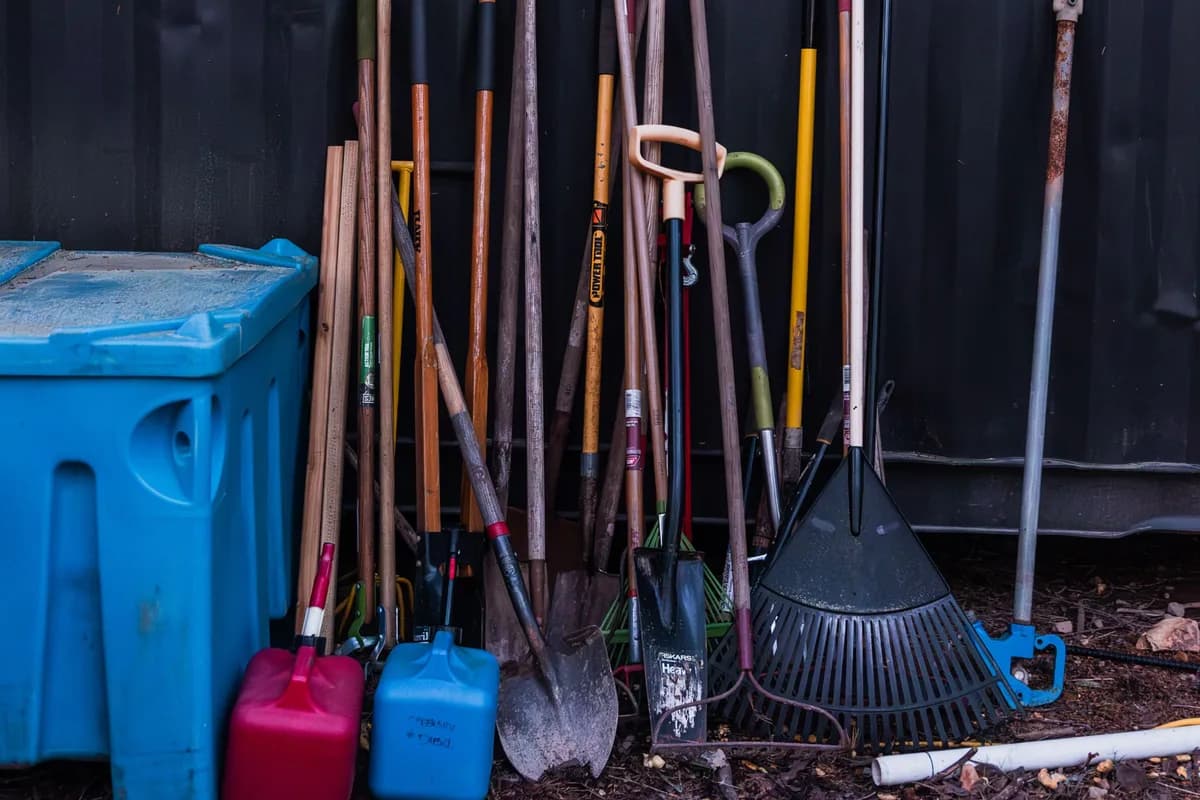 Spades, rakes and long-handled garden tools leaning against a dark metal wall beside a blue storage bin and two plastic fuel cans.