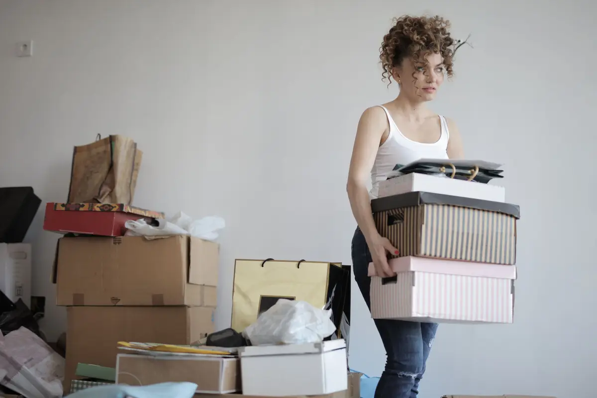 A woman carrying a stack of lidded boxes away from a pile of bags, cartons and loose items gathered on a bare floor.