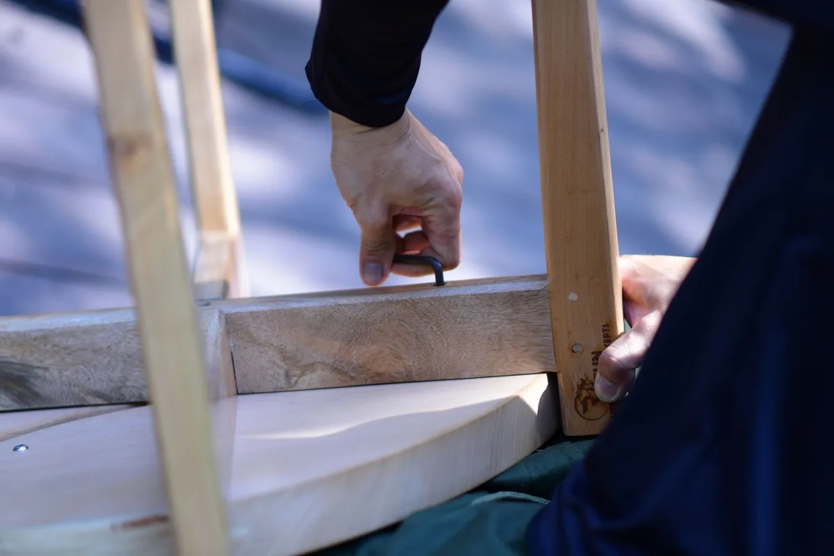 A person using a hex key to loosen a bolt on the joint of a light wooden furniture frame.