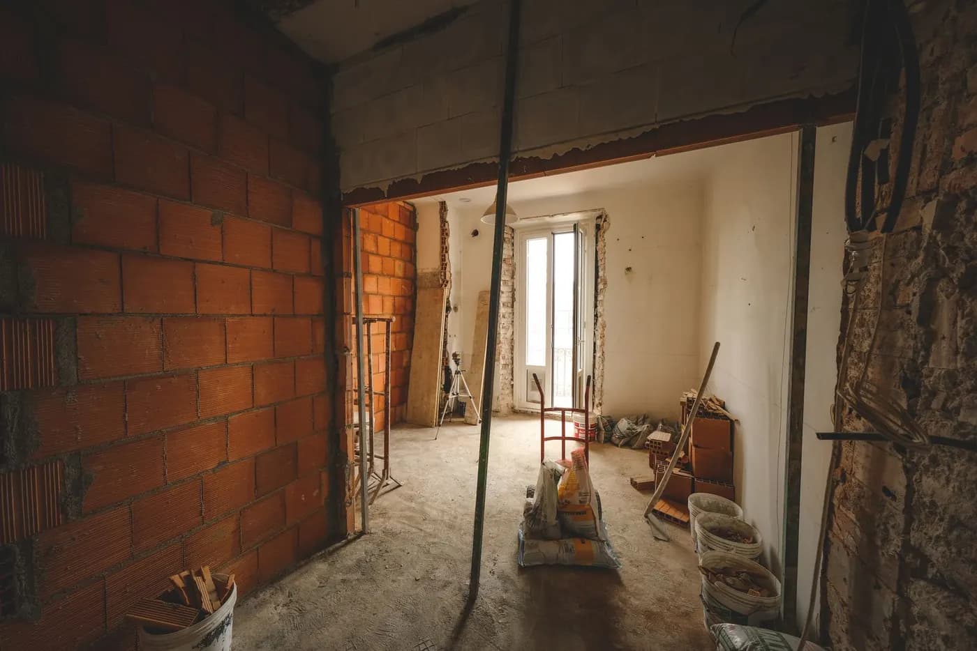 A room mid-renovation with exposed brickwork, buckets of rubble, stacked cement bags and support props on a bare concrete floor.