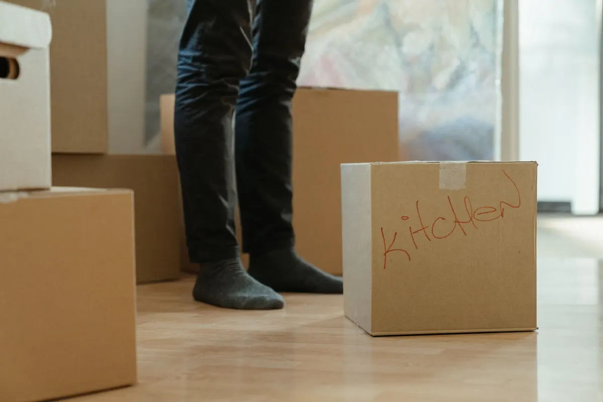 A cardboard box hand-labelled Kitchen in red marker on a wooden floor, with a person standing among more packed boxes.