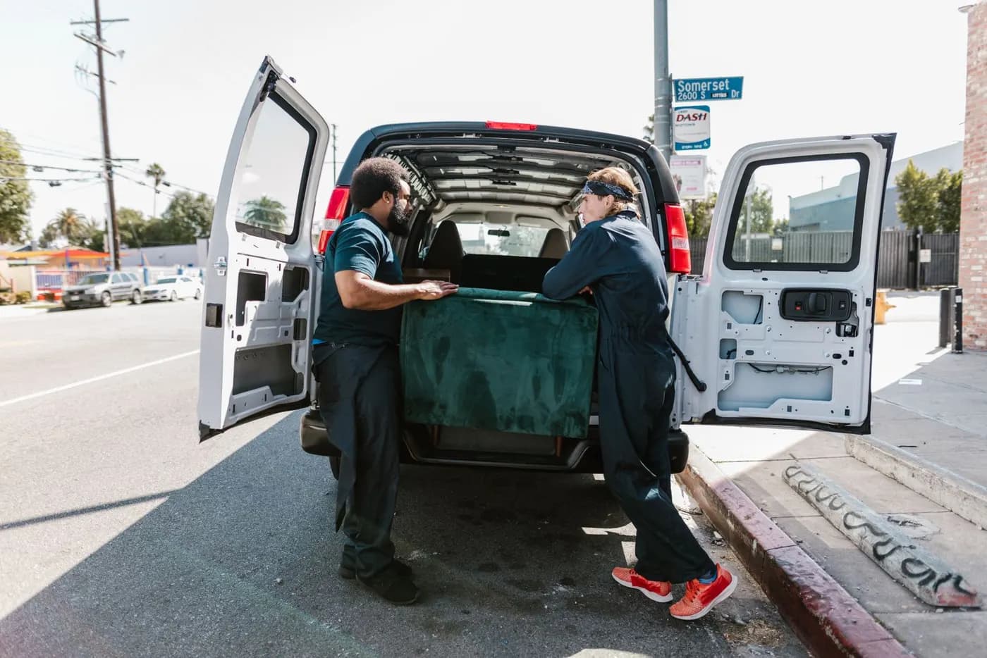Two removal workers loading a large sofa into the back of a van parked on a residential street.