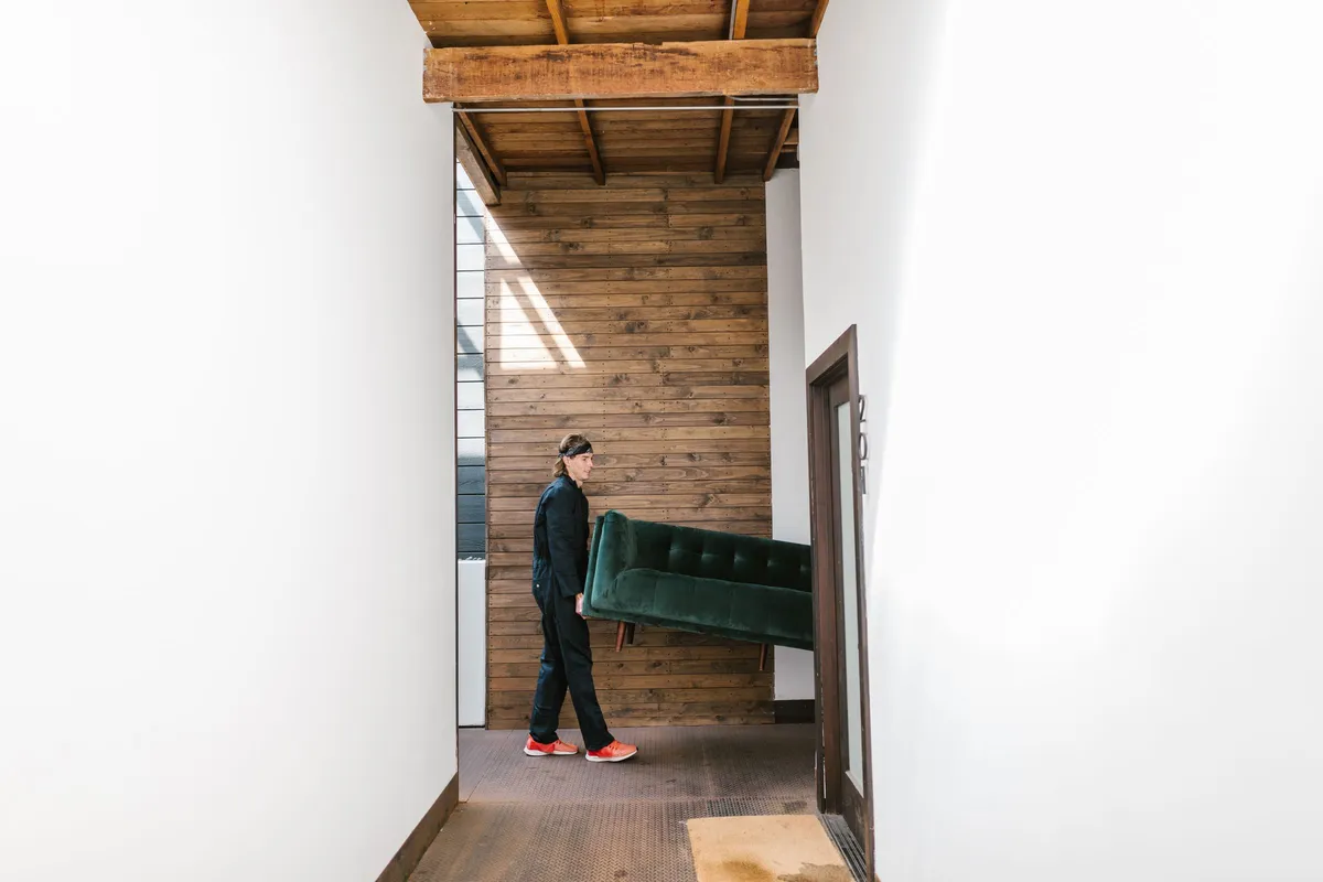 A mover in dark overalls carrying a green sofa on his own through a narrow white hallway towards an open doorway.