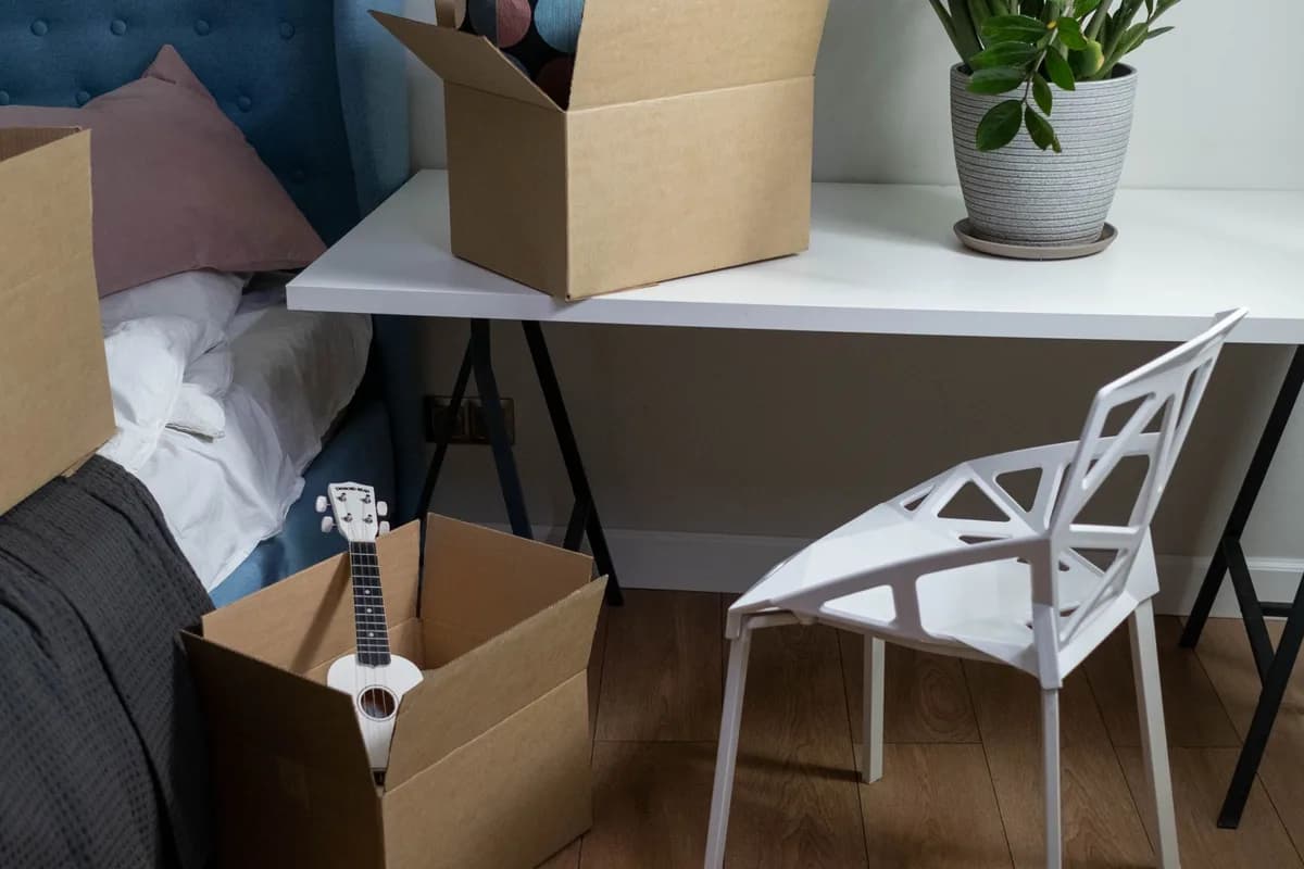 Open cardboard cartons part-filled with belongings on a bedroom floor, beside a white chair, a desk and a made bed.