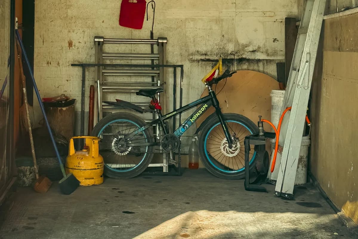 The inside of a concrete outdoor store holding a bicycle, a yellow gas cylinder, a folded ladder, buckets and a broom.