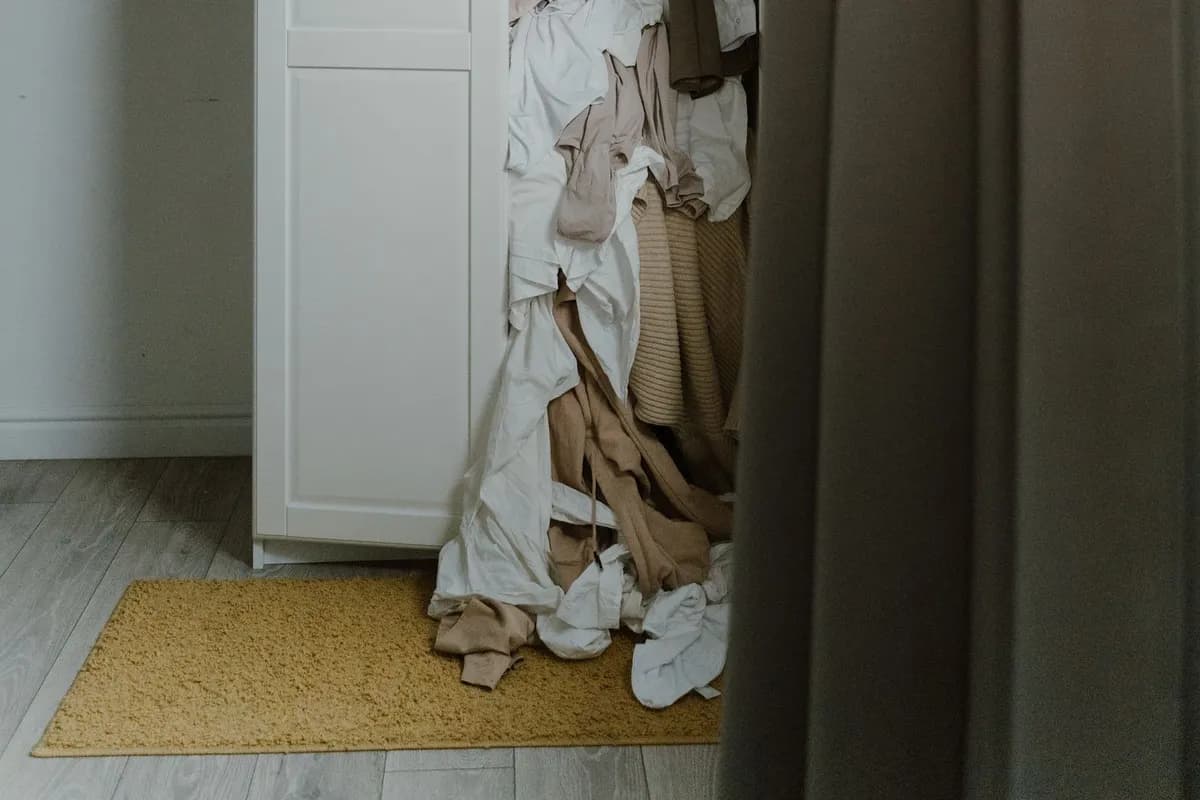 An open white wardrobe with clothes and linen spilling out onto a rug and wooden floor in a bedroom.