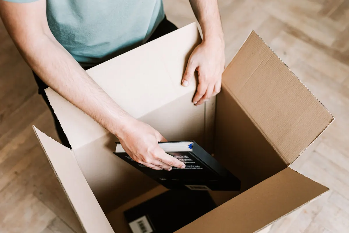 A person lowering hardback books into a cardboard box on a wooden floor.