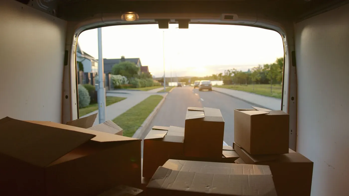 View from inside a part-loaded van looking out past stacked cardboard boxes onto a quiet residential street at sunset.