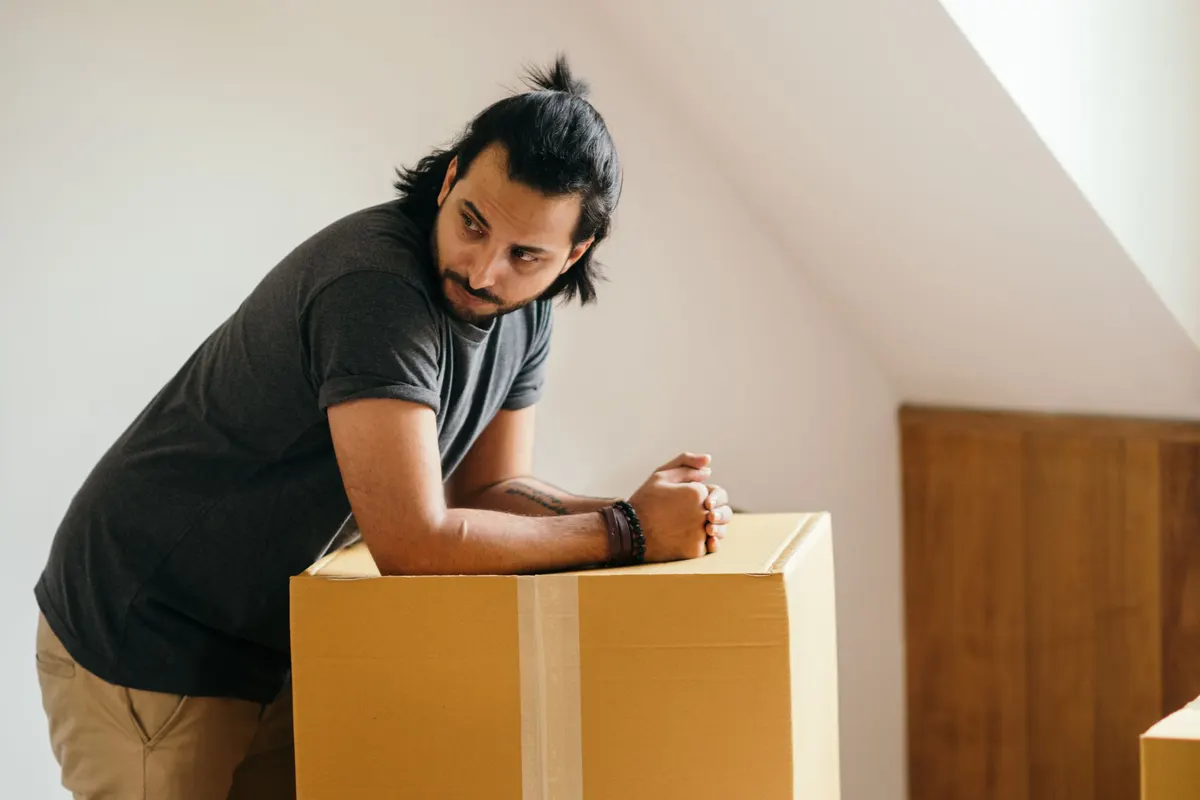 A man leaning on the top of a large taped cardboard box, pausing mid-way through clearing a room.