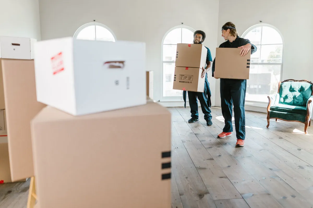 Two removal workers in dark uniforms carrying cardboard boxes across a bright, nearly empty room with tall arched windows.