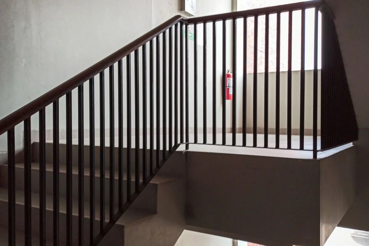 A concrete stairwell in a residential building with a dark metal handrail and a red fire extinguisher mounted on the landing wall.
