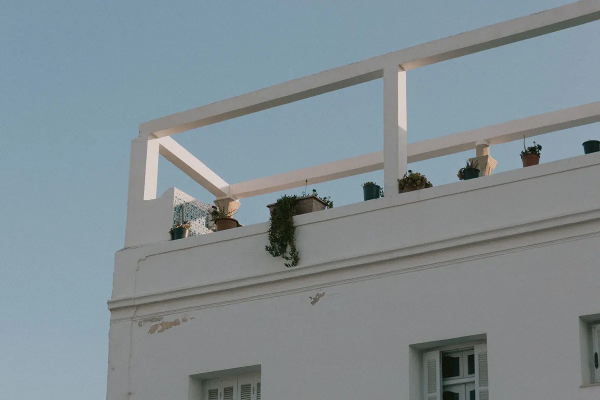 The white parapet and pergola frame of a roof terrace seen from below, with small potted plants lined up along the edge against a clear sky.