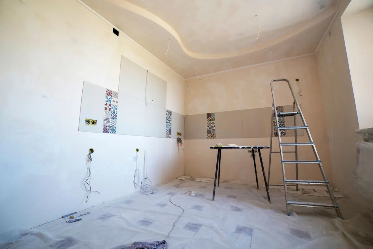 A room mid-renovation with dust sheets across the floor, a stepladder, exposed wiring and tile samples fixed to a half-finished wall.