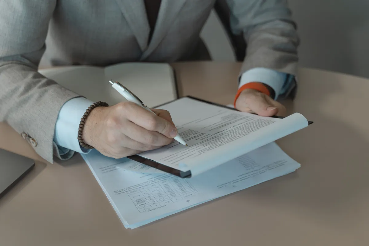 A close-up of someone reviewing and signing a printed document on a clipboard at a desk.