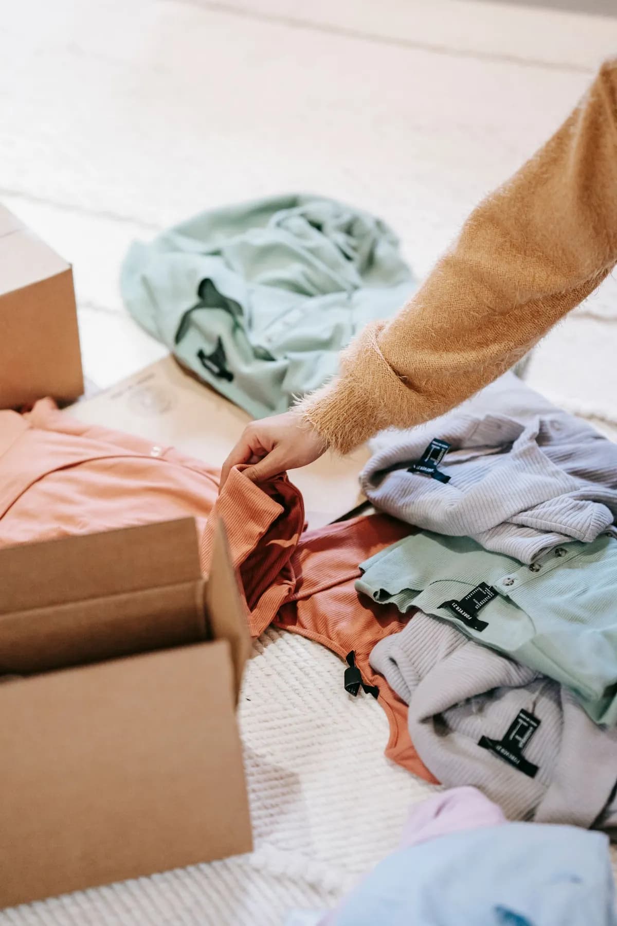 A person sorting folded clothes into a cardboard box next to other packed items on the floor.
