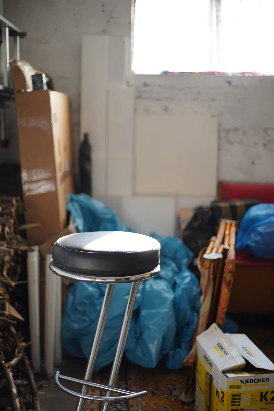 A cluttered storeroom with blue rubbish bags, flat-packed boxes, a bar stool and an appliance carton stacked against a bare wall.