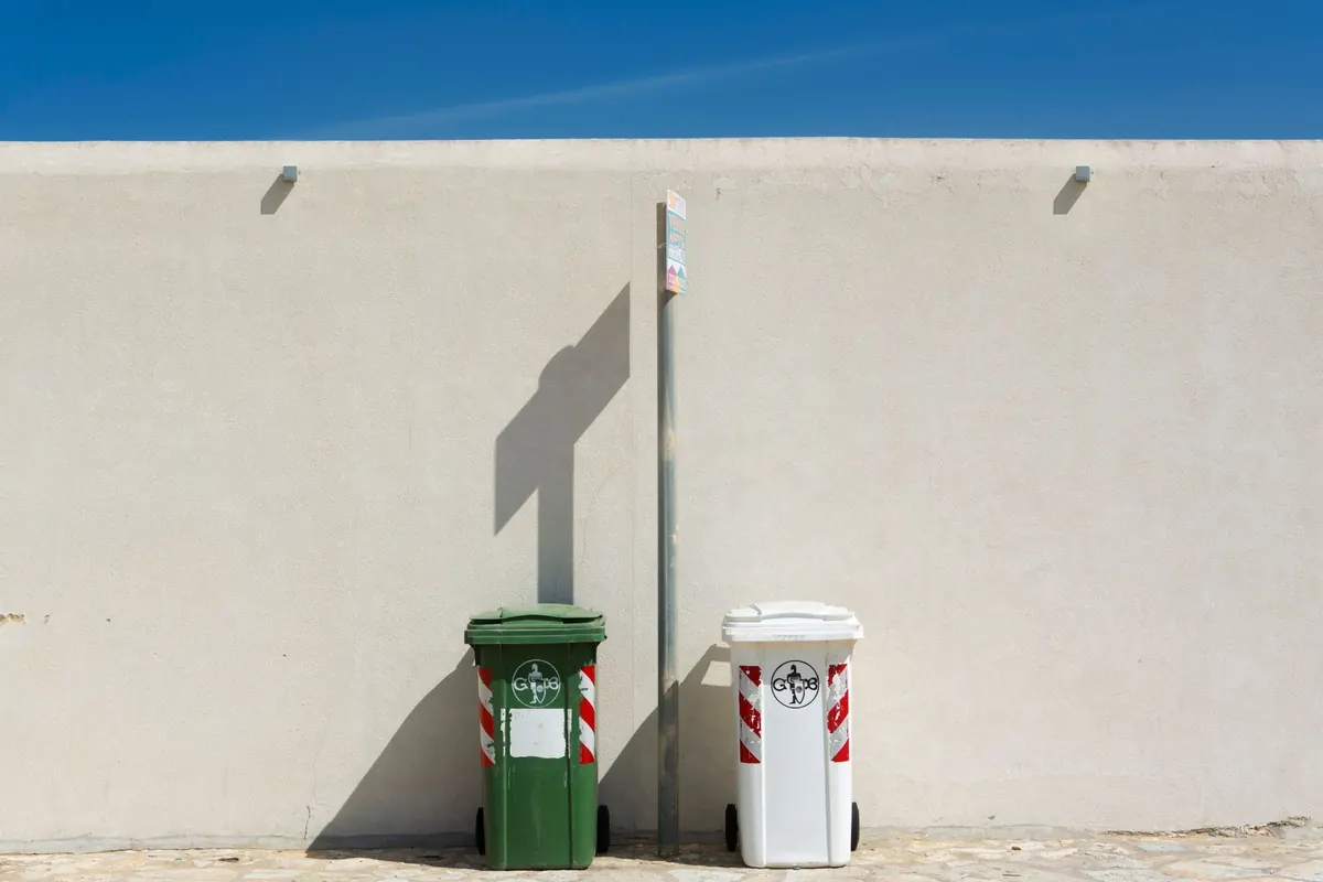 A green wheelie bin and a white wheelie bin standing against a pale plaster boundary wall in strong sunlight.