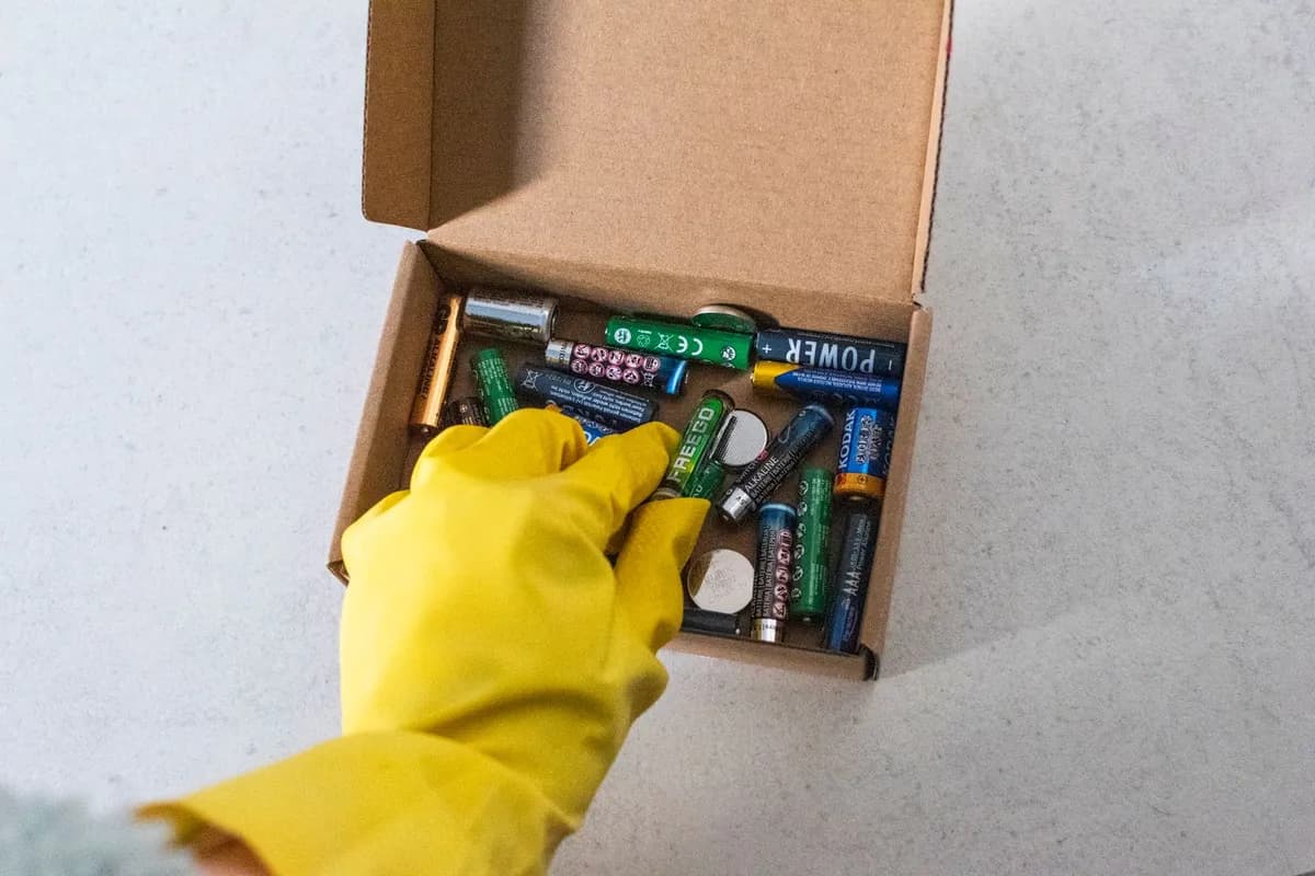 A gloved hand sorting a small box of used household batteries of different sizes and brands.