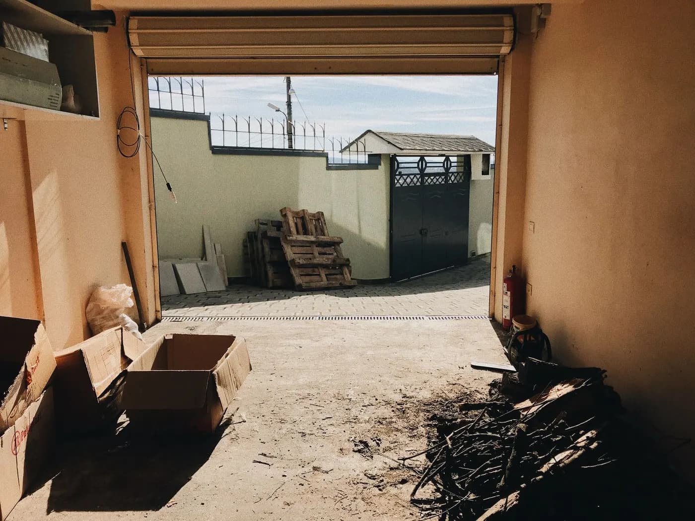 The interior of a villa garage looking out through an open roller door at a stack of wooden pallets and boxes in the courtyard.