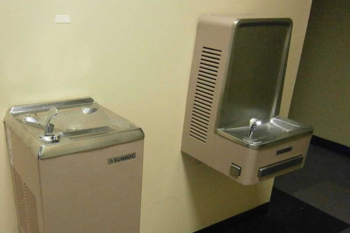 Two stainless steel drinking water coolers, one floor-standing and one wall-mounted, fixed to a cream wall in an office corridor
